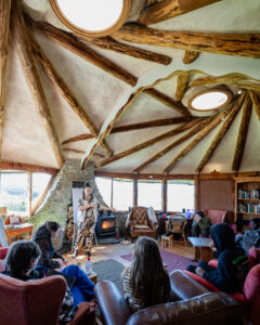 A photograph taken in the Hobbit Hut. Debris Stevenson addresses a group of people who are sitting in armchairs positioned in a circle. She is standing in front of a lit fire and a flipchart. Image captured by @smphotography.7 [Shaun Matheson]