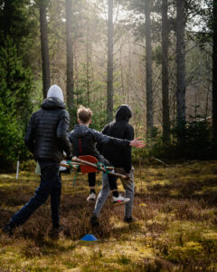 A photograph taken in Abriachan Forest. Two people in hoodies carry a young person on a homemade wooden raft through the trees. The person being carried sits facing their direction of travel, arms in the air. Image captured by @smphotography.7 [Shaun Matheson]