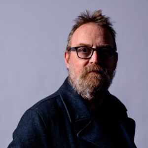 Boo's headshot. Boo is a white man with dark, thick rectangular glasses and a brown and grey beard. He is looking straight ahead, and wearing a dark blue denim jacket/blazer in front of a plain grey/blue coloured backdrop. His professional biography is adjacent.