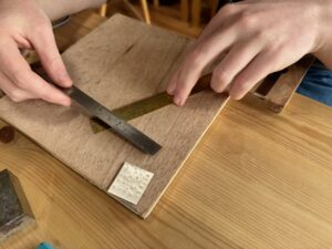 A photograph of a pair of hands using a metal ruler to measure the width of a piece of brass. They are leaning on a square block of wood. Image taken during a jewellery making workshop with Ailsa Wilde.