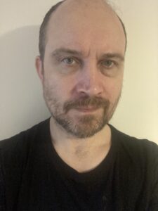 Chris' headshot. Chris is a white man with a short, dark beard and short hair. He is wearing a black round-neck top and is standing, looking straight ahead, in front of a beige background. His professional biography is adjacent.