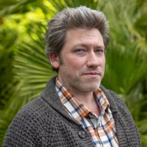 Luke's headshot. He is a white man with straight, dark hair. The background is made up of plants- mostly with long, thin green leaves. Luke is wearing a white, blue and orange checked shirt and a grey cardigan and is smiling towards the camera.