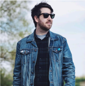 Mark's headshot. He is a white man with short black hair. Mark is wearing sunglasses, a navy and green checked shirt and a blue denim jacket. he is outside, looking into the distance on the right. His professional biography is adjacent.