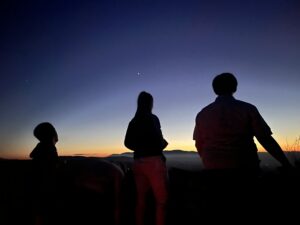 A photograph of 3 silhouettes looking out at the sunset. The sky melts, top to bottom, from navy, to pale blue, to a burning orange.