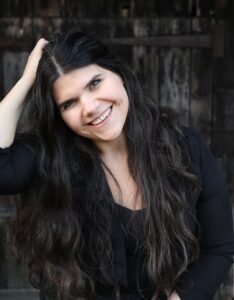 Michele's headshot. Michele is a white woman with long, wavy black hair, dark features and wearing a black top. She is smiling widely, leaning her head on her hand to the left. Behind her is what appears to be a wooden gate. Her bio is adjacent.